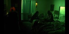 two people sitting on a bed in a room with green light coming from the window