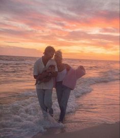 a man and woman walking on the beach with a baby in their arms at sunset