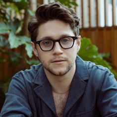 a young man wearing glasses sitting in front of a wooden fence with his arms crossed