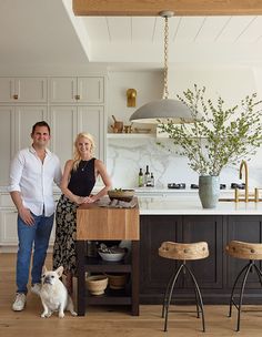 a man and woman standing in a kitchen next to a dog