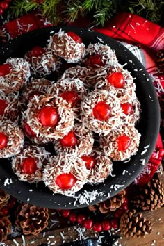 a black plate topped with cookies covered in chocolate and cherries next to pine cones