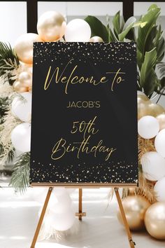 a black and gold welcome sign sitting on top of a wooden easel next to balloons