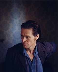 a man in a blue shirt and black jacket looking off into the distance with his hand on his head