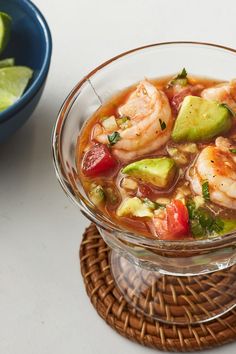 a bowl of soup with shrimp, avocado and tomatoes on the side next to a basket of lime wedges