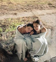 two women wrapped up in blankets sitting on the ground next to an open field and dirt road