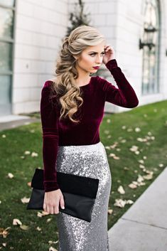 a woman with long hair wearing a burgundy top and silver sequin skirt, standing in front of a building