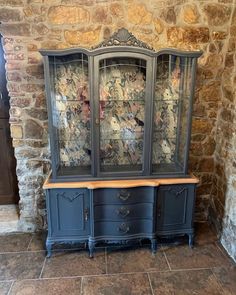 an old china cabinet with glass doors in a stone walled room, next to a brick wall