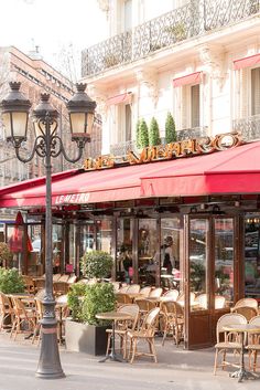 an outdoor cafe with tables and chairs under a red awning next to a street lamp