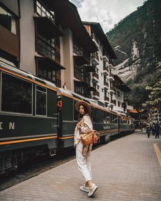 a woman is standing in front of a train at a station with mountains in the background