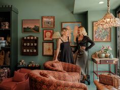 two women standing next to each other in a room with pictures on the wall and furniture