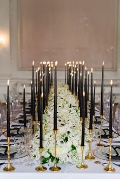 a long table with black candles and white flowers on it is set for a formal dinner