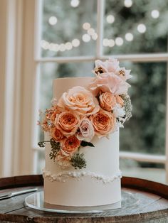 a white wedding cake with pink flowers and greenery on top sits in front of a window