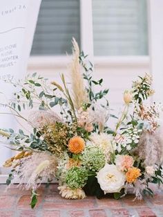 an arrangement of flowers and greenery sits on a brick floor next to a sign