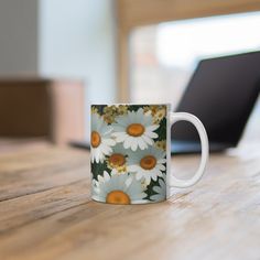 a coffee mug with daisies on it sitting on a table next to a laptop