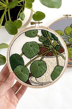 a hand is holding two embroidery hoops with green leaves on them and one has a potted plant in it