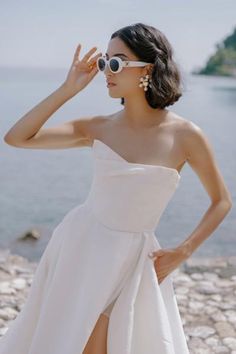 a woman in a white dress is standing on the rocks by the water wearing sunglasses