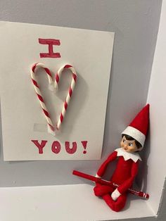 an elf is sitting on the shelf next to a valentine's card and candy cane