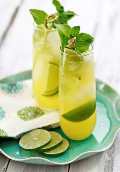 two glasses filled with lemonade and cucumber on a green plate next to lime slices