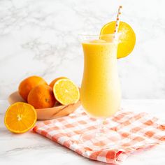 an orange smoothie in a glass next to some oranges on a checkered cloth