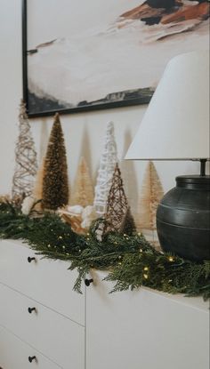 a christmas mantle with pine cones and evergreen garland on it, next to a lamp