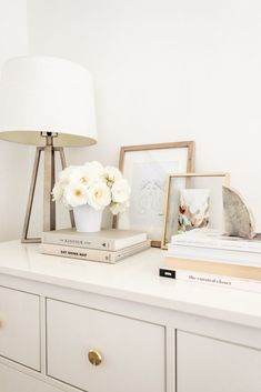 a white dresser with flowers and books on it next to a lamp in a bedroom