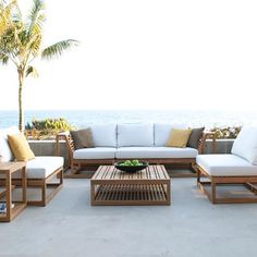 a couch and table sitting on top of a cement floor next to the ocean with palm trees in the background