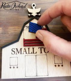 a hand is holding a small wooden house with an american flag on it and the words small town written in red white and blue