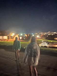 two people walking down a path at night with lights on the buildings in the background