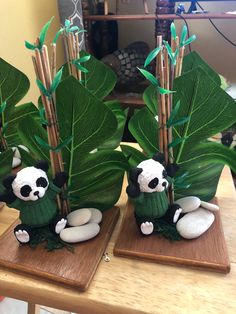 two small stuffed panda bears sitting on top of wooden bases next to green plants and bamboo sticks