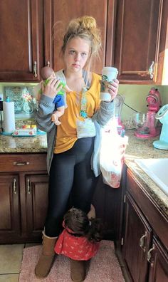 a woman standing next to a child in a kitchen holding two cups and looking at the camera