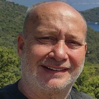 an older man smiling for the camera in front of some trees and mountains with a body of water behind him