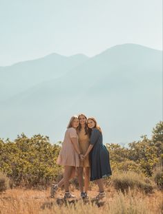 three women are standing together in the grass with mountains in the background