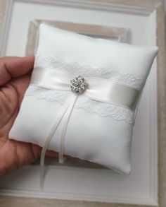 a hand holding a white ring pillow with a ribbon and jewel brooch on it