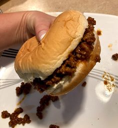 a hand holding a sloppy joe sandwich over a white plate