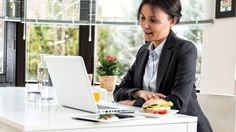 a woman sitting in front of a laptop computer eating a sandwich and drinking orange juice