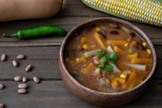 a wooden bowl filled with soup next to corn on the cob and green peppers