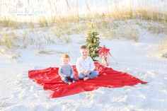 two young boys sitting on a blanket in the sand with a christmas tree behind them