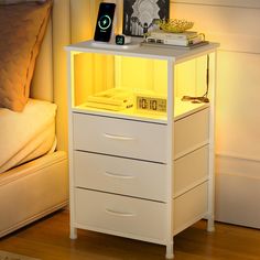 a white night stand with two drawers and an alarm clock on the top, next to a bed