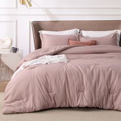 a bed with pink comforter and pillows in a white room next to a lamp