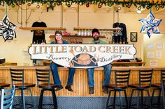 two people sitting at a bar holding a sign that says little toad creek brewery and bakery