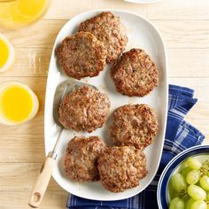 some meat patties are on a white plate with grapes and orange juice in the background