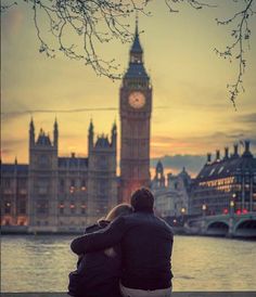 two people sitting on a bench looking at the water and clock tower in the background