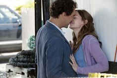 a man and woman kissing each other in front of a white wall with cars behind them