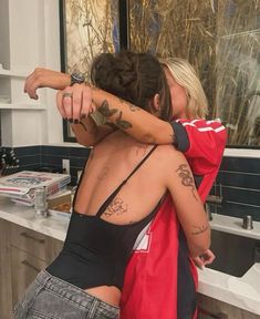 two women hugging each other in front of a bathroom sink and counter top with tattoos on their arms