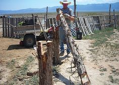 a man standing next to a wooden fence in the middle of a dirt field with mountains in the background