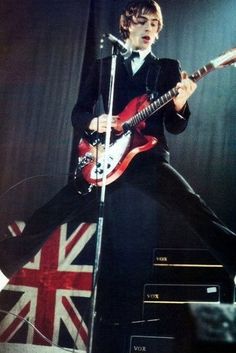 a man in a tuxedo playing an electric guitar on stage with the british flag behind him