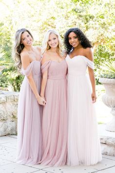 three bridesmaids pose for a photo in front of a fountain wearing off - the - shoulder gowns