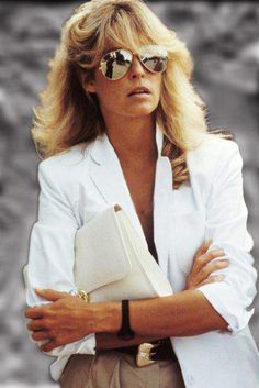 a woman wearing sunglasses and a white blazer with her arms crossed, looking off to the side