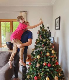 a man holding a child in his arms near a christmas tree with decorations on it