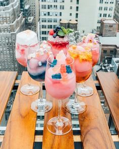 four glasses filled with different types of drinks on top of a wooden table in front of tall buildings